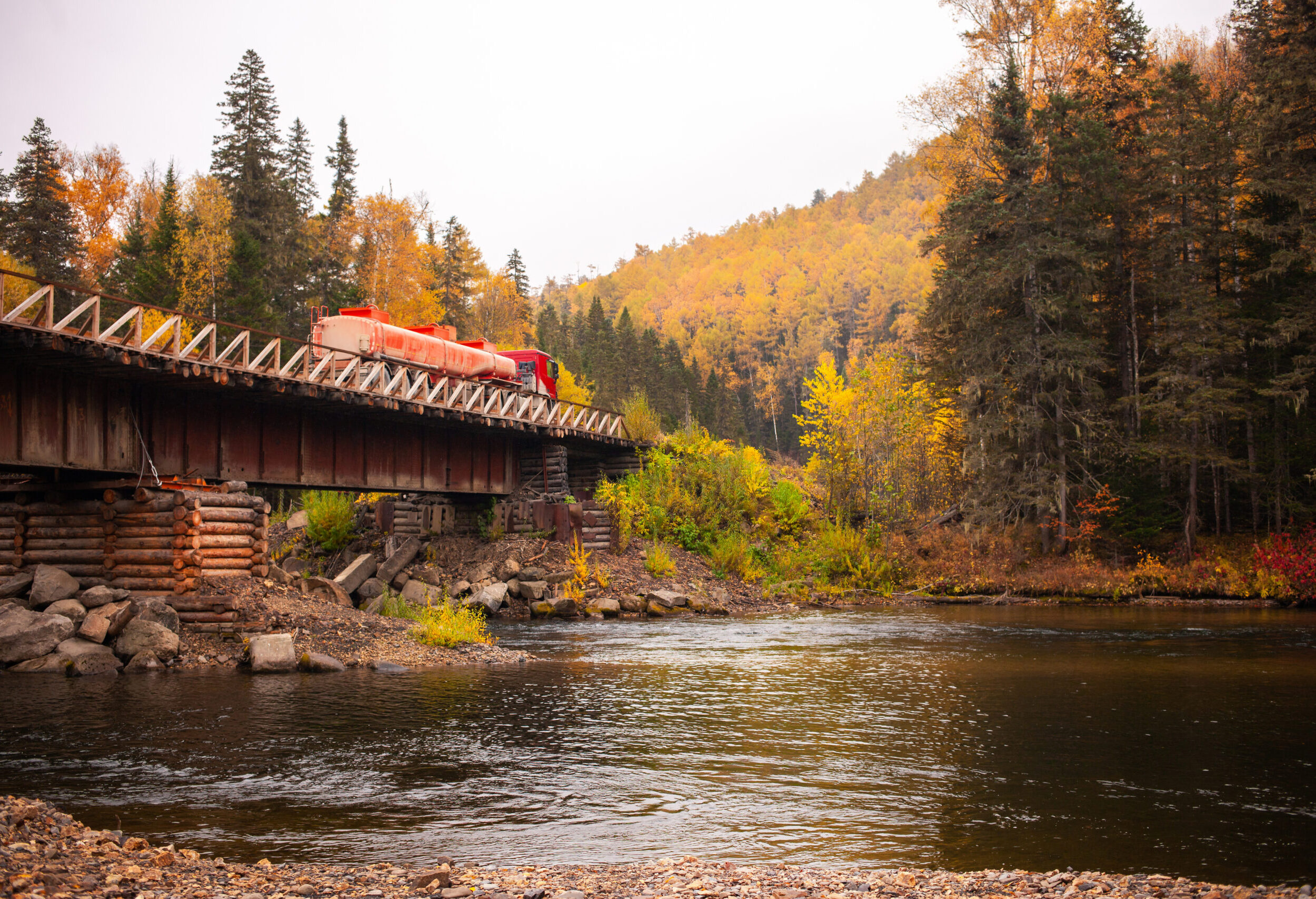 Red fuel truck at wooden bridge over mountain river in autumn